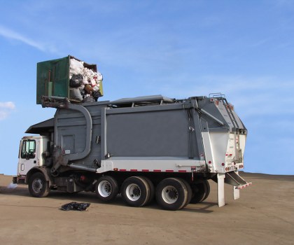 A large commercial waste collection truck parked on a flat, paved surface under a clear blue sky. The truck has a white cab with a small window, and its main body is covered with a dark grey, weathered metal hopper designed for waste storage and loading. The rear section features a hydraulic lifting mechanism with a hinged panel for unloading rubbish, and there is a small pile of black waste bags lying on the ground nearby. At the top of the truck's hopper, an open green bin is visible, overflowing with mixed waste including crumpled paper, plastic bottles, and cardboard, some of which are spilling out. The scene suggests a rubbish collection operation, consistent with services offered by Commercial Waste Potters Bar for local businesses in the area, possibly within the local postcode region or nearby towns.