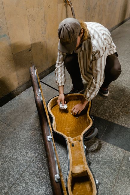 A person wearing a gray cap, striped shirt, and dark pants is crouched down on a tiled floor, placing money into an open, brown, vintage-style guitar case with a plush interior. The guitar case is lyi