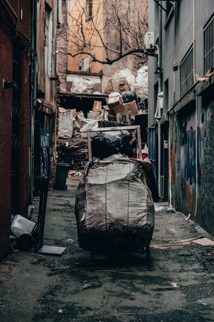 A person wearing a blue and yellow checkered shirt, grey trousers, and light green gloves is holding open a large black rubbish bag, preparing for waste disposal outdoors. The bag is made of glossy pl