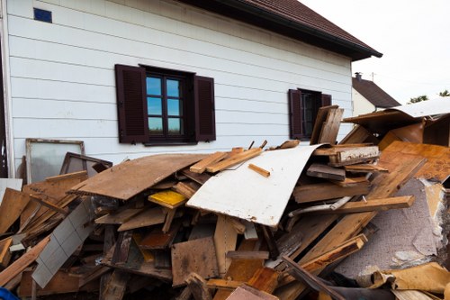 A large pile of discarded wooden planks, broken furniture parts, and various construction debris is stacked against the white exterior wall of a residential building in Potters Bar, Hertfordshire, near the driveway. The timber pieces vary in size, thickness, and finish, with some showing chipped edges and weathered surfaces, predominantly in natural brown tones. Among the debris, there are also a few white panels and a yellow plastic item partially visible. The building features dark brown window shutters, with two windows in view, and a dark tiled roof extending above the wall. The surrounding environment includes a paved driveway and other residential rooftops in the background, suggesting an urban or suburban setting. The scene is illuminated by natural daylight, giving a clear view of the details, consistent with waste collection or rubbish removal activities that a company like Commercial Waste Potters Bar might handle, especially in the context of property clearance or renovation waste management services.